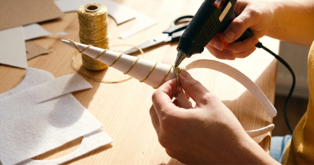 Hands making a DIY unicorn horn by wrapping a cardstock cone in twine and gluing it to a headband